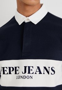 Polo navy con cuello blanco en contraste y franja horizontal blanca que presenta el texto "PEPE JEANS LONDON"; material de algodón, corte relajado.