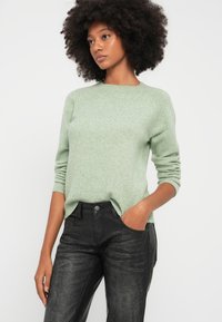 Vaaleanvihreä neulepaita, jossa on pyöreä pääntie ja pitkät hihat, yhdistettynä mustiin teksturoiduihin housuihin, joissa on sivutaskut ja kevyt kulutus.