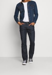 Jeansjacka i mörkblått med ljusbrun krage, i kombination med mörk denimjeans och svarta canvasmärkeskor, stående mot en enfärgad bakgrund.