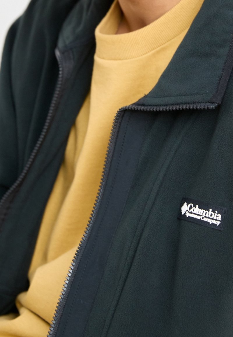 Mörkgrön fleecejacka med dragkedja framtill, med en svart Columbia-logotyp på vänster bröst, bärs över en ljusgul sweatshirt.