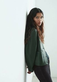 Giacca verde realizzata in un materiale morbido con una vestibilità rilassata, dotata di due tasche frontali, abbinata a jeans scuri. Il modello si appoggia a un muro.