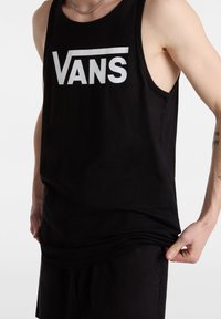 Personne portant un débardeur noir avec un logo "VANS" blanc, tenant l'ourlet inférieur, montrant une partie de ses tatouages sur le bras.
