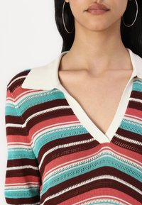 Polo en tricot rayé avec un col en V, arborant des bandes horizontales en teal, rose, bordeaux et blanc. Fil texturé et col contrastant.