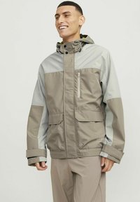 Kurtka GORE-TEX w beżowo-szarym schemacie kolorystycznym. Posiada zapinaną na zamek kieszeń na piersi, przednie klapy oraz regulowany kaptur, ukazując gładką fakturę.