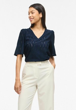 Kobieta ubrana w granatową, teksturowaną bluzkę z dekoltem w serek i kremowe spodnie z wysokim stanem, uśmiechająca się i patrząca w bok, z jedną ręką w kieszeni.