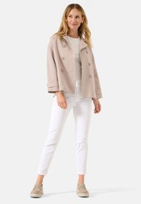 Hellbeige Leinenjacke mit lockerer Passform, doppelten Knopfverschlüssen und Rundkragen, kombiniert mit weißen schmalen Hosen und beigen Slippern.