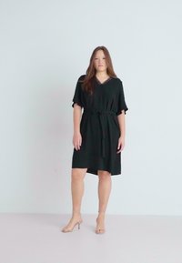 Black dress with short flutter sleeves, V-neck, belted waist, and knee-length hem. Smooth fabric with subtle sheen. Worn with nude heeled sandals.