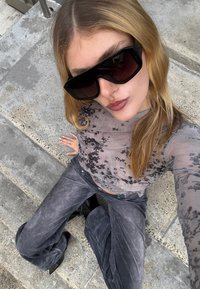 Prozirni sivi top s cvjetnim uzorkom, sive baršunaste hlače i velike crne sunčane naočale. Glatka tekstura s pripijenim krojem. Vidljive crne čizme.