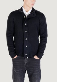 Uomo che indossa un cardigan nero abbottonato sopra una camicia bianca, con le mani nelle tasche di jeans neri sbiaditi, su uno sfondo chiaro.