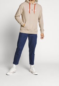 Sudadera beige con capucha, cordones naranjas y logo blanco, combinada con pantalones azul marino y zapatillas altas blancas.