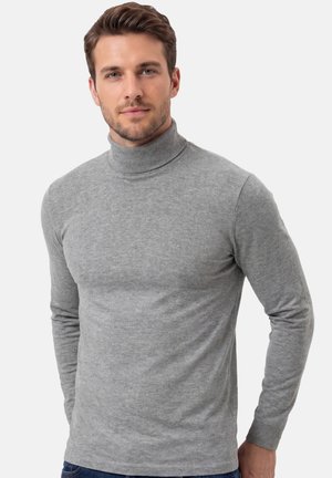 Mann mit kurzem braunem Haar, der einen eng anliegenden hellgrauen Rollkragenpullover trägt und vor einem schlichten Hintergrund steht.