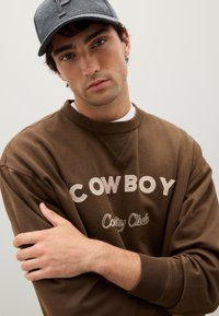 Sweatshirt marrone con la scritta "COWBOY" e "Cozy Club" ricamate in crema. Tessuto morbido, collo rotondo e vestibilità rilassata; indossato con un berretto grigio.
