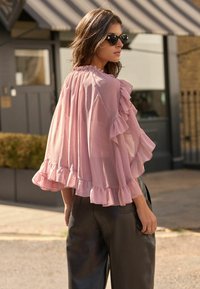 Durchsichtige rosa Bluse mit Rüschensleeves und Saum, kombiniert mit hochgeschnittenen schwarzen Hosen. Accessoires umfassen runde schwarze Sonnenbrillen.
