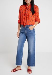 Mujer vestida con una blusa floral naranja con un lazo, jeans de pierna ancha azules, sandalias rojas y un reloj, de pie con una mano en el bolsillo.