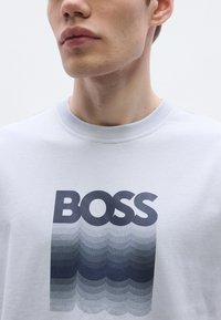 Helehall tooniga pusa, millel on trükitud tumedat halli värvi logo „BOSS” all laineline gradientmuster. Soonikkoes kaelus.