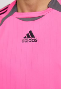 Adidas rožinė sporto marškinėliai su vertikaliomis juostelėmis, apvalia iškirpte ir juodu logotipu. Akcentai yra pilki pečių srityje, pagaminti iš tekstūruotos medžiagos.