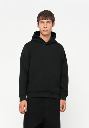 Junger Mann in einem schwarzen Kapuzenpullover mit einer Fronttasche, der vor einem schlichten weißen Hintergrund steht.