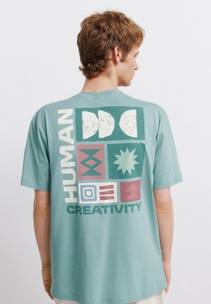 Młody mężczyzna z krótkimi brązowymi włosami, ubrany w seledynowy t-shirt z geometrycznymi kształtami i napisami "HUMAN CREATIVITY" na plecach.