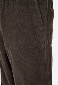 Brūni korduroja bikses ar vertikālām ribām, ar sānu kabatām un izšūtu logotipa detaļu kontrastējošā diegu krāsā.