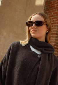 Dunkelgrauer Wollmischungs-Mantel mit hohem Kragen und zwei Knöpfen, kombiniert mit einer Schildpatt-Sonnenbrille und einem passenden grauen Schal.