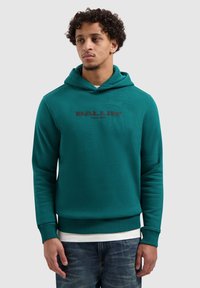 Petrolblauwe sweatvest met capuchon, voorzien van een zwart "BALLIN"-logo over de borst, geribbelde manchetten en een gladde textuur. Gedragen over een wit overhemd.