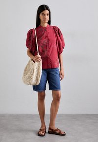 Rote Bluse mit Blumenmuster und Puffärmeln, kombiniert mit Denim-Shorts. Das Model trägt eine cremefarbene geflochtene Tasche und braune Sandalen.