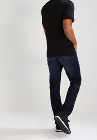Unos jeans de mezclilla oscura con un corte ajustado, combinados con una camiseta negra de manga corta y zapatillas deportivas negras, mostrados desde atrás.