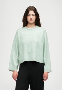 Un pull court vert menthe avec un col rond, des épaules tombantes et des poignets côtelés. Associé à un pantalon noir à jambes larges.