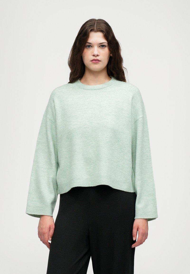 Un pull court vert menthe avec un col rond, des épaules tombantes et des poignets côtelés. Associé à un pantalon noir à jambes larges.