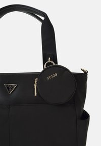 Černá nylonová taška s pevným designem, obsahuje zlaté trojúhelníkové logo a odnímatelnou kulatou peněženku na mince. Hladký povrch a pevné rukojeti.