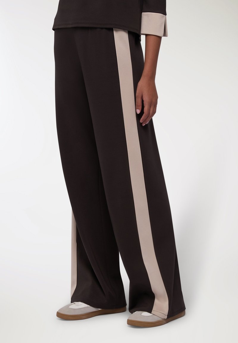 Pantaloni neri a gamba larga con strisce laterali beige, realizzati in un tessuto morbido. Abbinati a scarpe grigie chiare e beige, con un design casual.