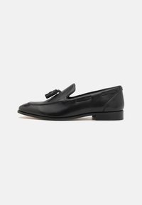 Mocasines de cuero negro con puntera redondeada, acento decorativo de borlas y detalles recortados en los laterales; tacón plano y textura suave.