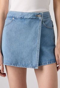 Ljusblå denim minikjol med omslagdesign, en framficka och kontrasterande sömmar. Stängningar består av en knapp och dragkedja.