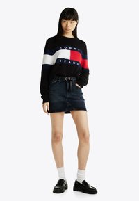 Maglione nero con strisce orizzontali rosse, bianche e blu, con la scritta "TOMMY JEANS", abbinato a una gonna di jeans scura e mocassini neri.