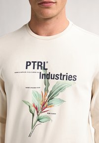Lichtbeige sweatshirt van katoen, met een bedrukt botanische illustratie en zwarte tekst voor "PTRL Industries" en locatiegegevens.