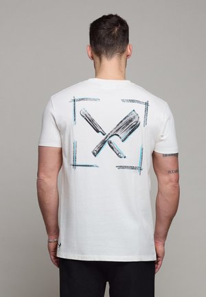 Mann iført hvit t-skjorte med svart og blåt bilde av en barberhøvel og kjøttkniv krysset på ryggen, stående mot en ensfarget bakgrunn.