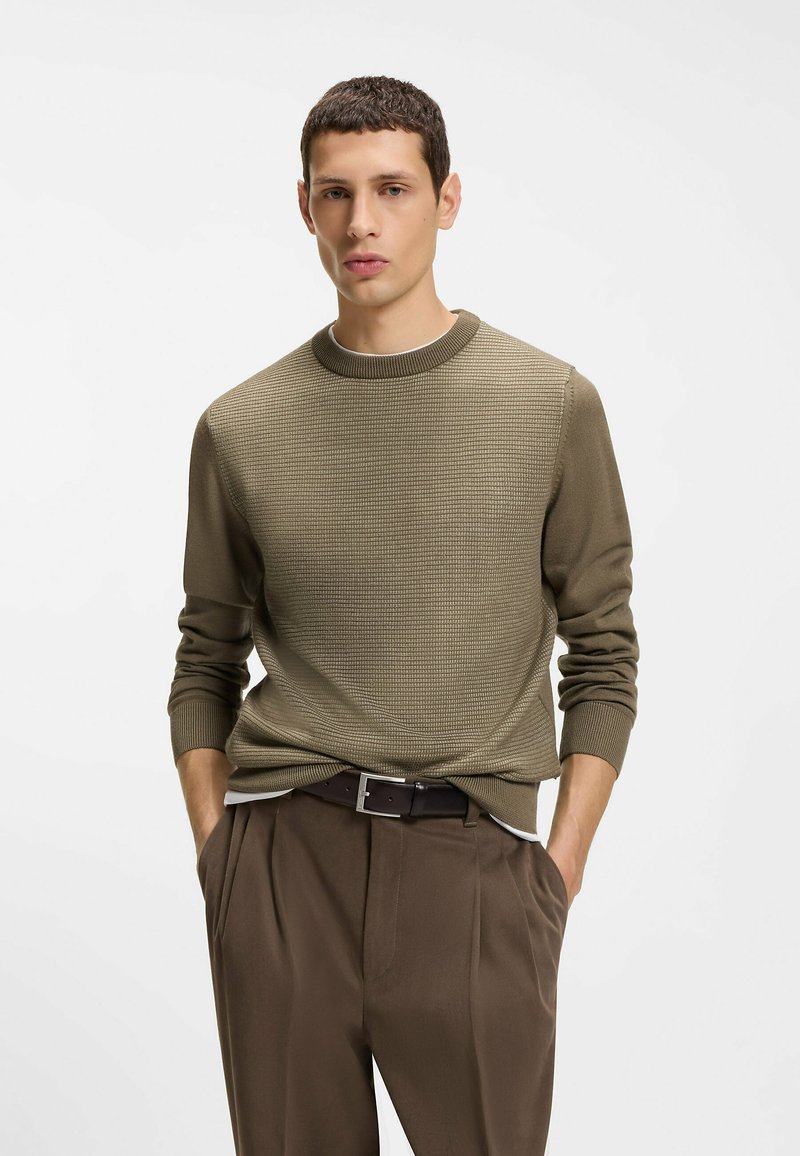 Pull texturé vert olive avec un col rond. Associé à un pantalon marron et une ceinture assortie, avec des poches latérales et des plis.