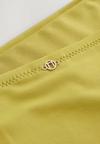 Limegrön tyg med ribbad textur; har en guldfärgad cirkulär hårdvaruaccent vid sömmen med dekorativ sömnad ovanför.