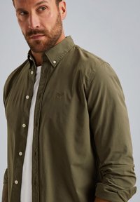 Camisa de botones color verde oliva con textura suave, mangas arremangadas y botones de color claro. Presenta un logo bordado en el pecho.