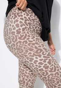 Osoba oblečená v béžových a hnědých legínách s leopardím vzorem a černém topu, jedna ruka opřená o bok na bílém pozadí.