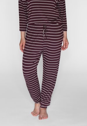 Personne portant un pantalon de pyjama ample violet foncé et un haut avec des rayures horizontales blanches, debout pieds nus sur un sol clair.