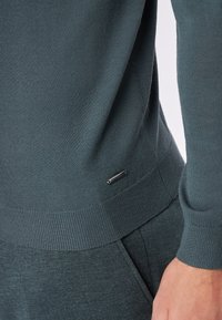 Dunkelblauer Pullover mit geripptem Muster, ausgestattet mit einem kleinen metallischen Logo-Element am Saum und eng anliegenden Bündchen. Der Stoff wirkt weich und glatt.