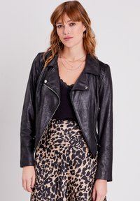 Veste de biker en faux cuir noir avec des fermetures éclaires et des pressions argentées, portée sur un haut noir, associée à une jupe beige à imprimé léopard.