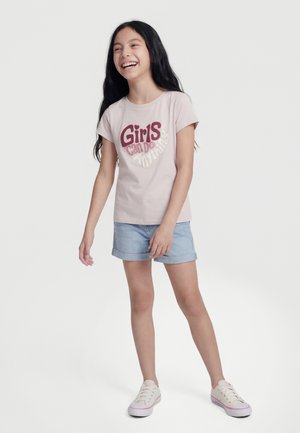 Smilende pige med langt mørkt hår iført en lyserød t-shirt med teksten "Girls can do anything", denimshorts og hvide sneakers stående på hvid baggrund.