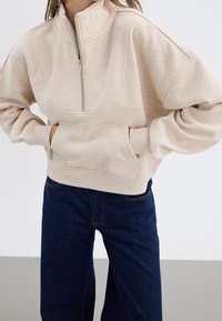 Ljusbeige halvdraget sweatshirt med en avslappnad passform, ribbade ärmmuddar och detaljerad framficka. Bärs med mörkblå raka jeans.
