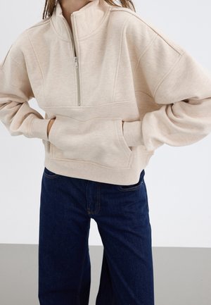 Licht beige half-zip sweatshirt met een relaxed fit, ribbelmanchetten en een voorzak detail. Draagt met donkerblauwe rechte jeans.