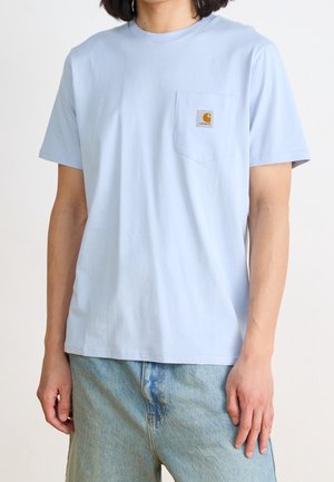 Osoba ubrana w jasnoniebieski T-shirt z krótkim rękawem, kieszenią z przodu i naszywką z logo, w połączeniu z jasnoniebieskimi, wypranymi jeansami.