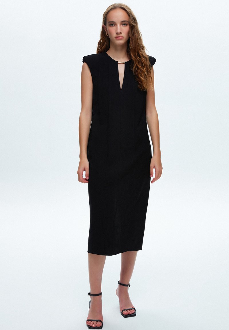 Vestido negro sin mangas con escote en V, hombros estructurados y longitud hasta la mitad de la pantorrilla. Tela suave con costuras verticales. Tacones negros con tiras.