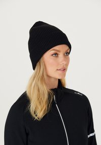 Sort strikket hue med ribbet tekstur og spidst top, båret af en person i en sort zip-up jakke med hvide detaljer.