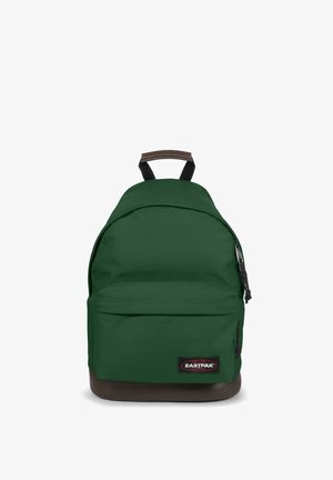 Mochila de tela verde con forma redondeada, base marrón, bolsillo frontal con cremallera y una etiqueta con el logo en negro. Correas ajustables negras.